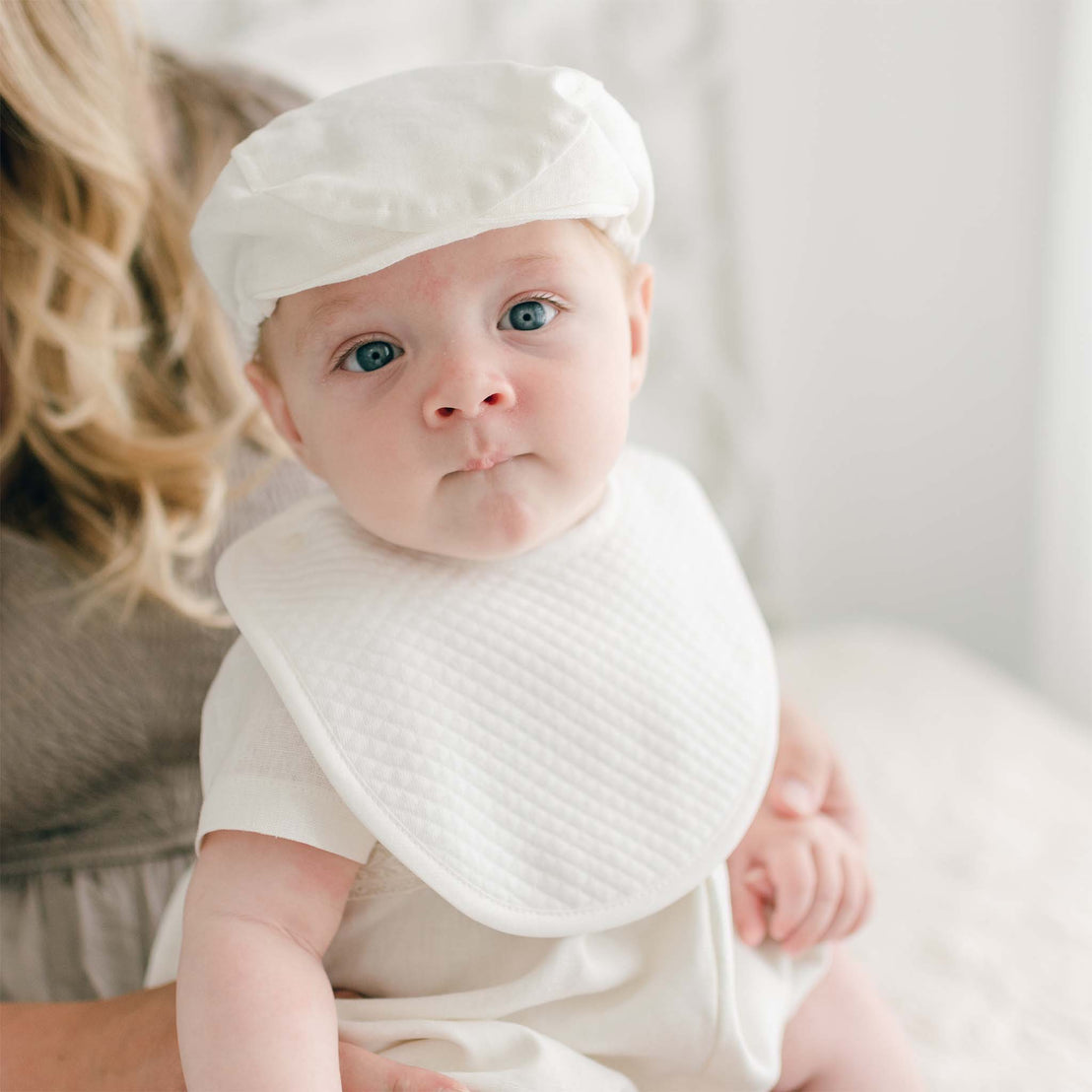 A baby wearing a white cap and an Oliver Bib with button closure sits on a woman's lap. The baby has a curious expression and is looking towards the camera. The background is softly lit and out of focus. The woman is partially visible, with only her hair and shoulder showing.