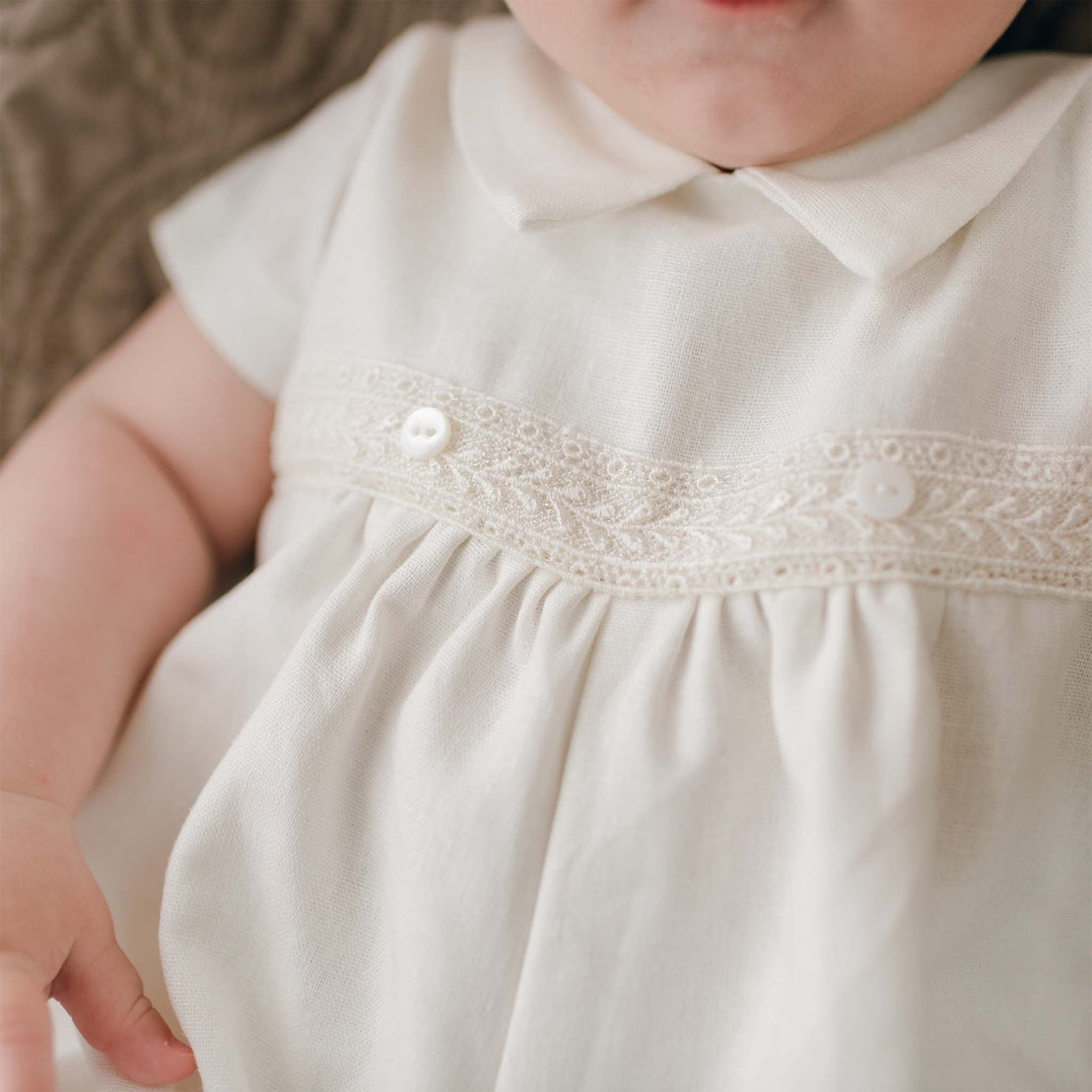 The details on the bodice of the Oliver Romper in ivory. A baby boy is wearing the romper. The details show lace that adorns the front bodice as well as buttons. 