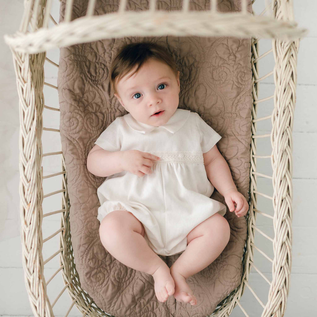 Baby boy wearing the Oliver Romper in ivory. He is laying in a wicker basket lined with a brown fleur-de-lys quilted pattern blanket. He looks up with a curious gaze.