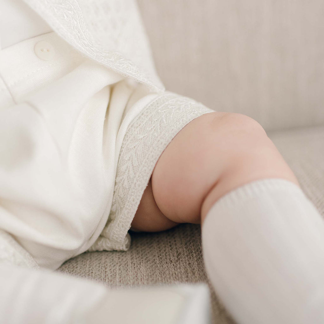 Close-up of a baby boy's chubby leg dressed in the light ivory Oliver Sweater Shorts that feature a delicate linen blend, accented with a pearl button and Venice lace along the hem. The setting is a soft, neutral-toned background.