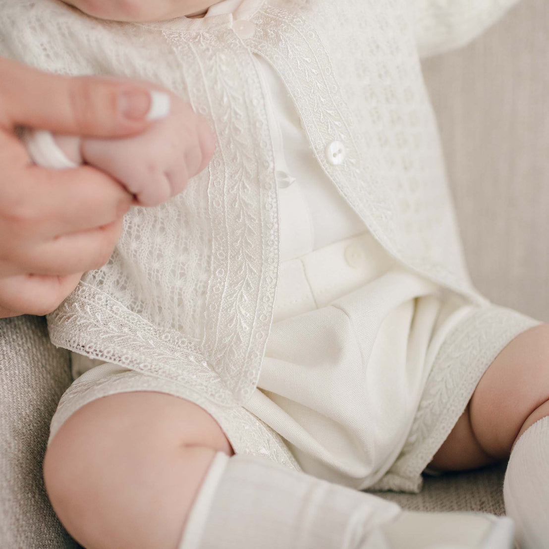 Close-up of a baby dressed in the light ivory Oliver Sweater Shorts Suit, holding an adult's finger. Only the Oliver Knit Sweater and Shorts are visible. The background is a soft, neutral-toned background.