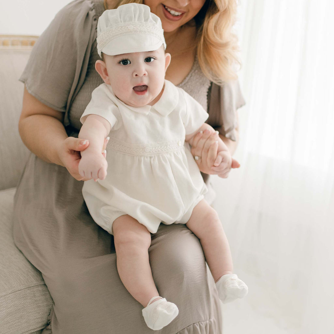 Baby boy sitting on chair with mom and wearing the Oliver Romper, a bubble romper made from linen featuring a collar and bodice with buttons and Venice lace detail (without the Oliver Convertible Gown).