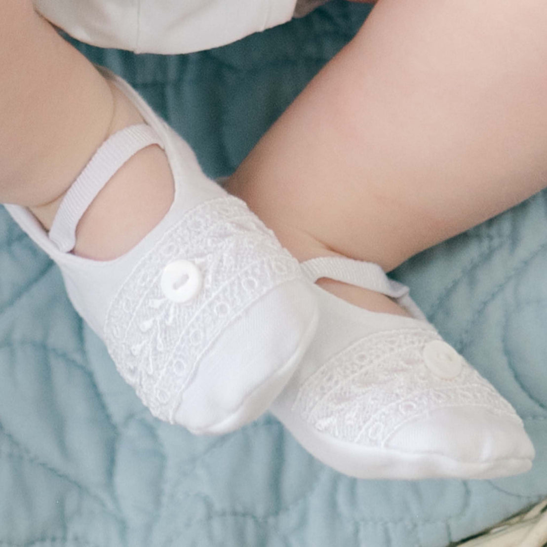 The top detail of the Oliver Booties in white worn by a baby boy. The Oliver booties are made with a linen top, soft pima cotton bottom, and features a Venice lace and button detail (with elastic strap across the foot). The background is a blue quilted blanket with fleur-de-lys design.