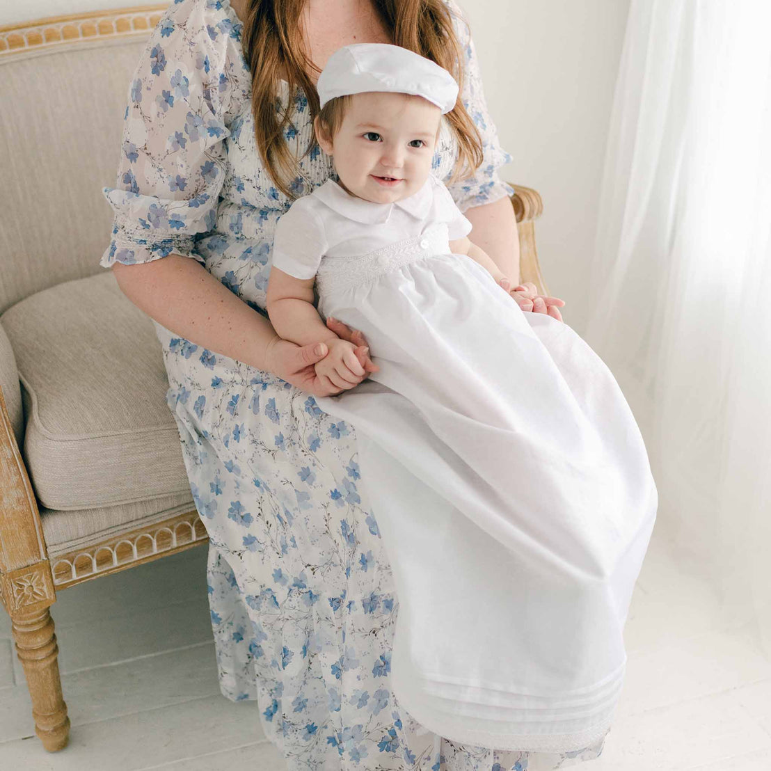 Baby boy sitting on a chair and wearing the White Oliver Convertible Gown and Romper (with matching Linen newsboy Hat).