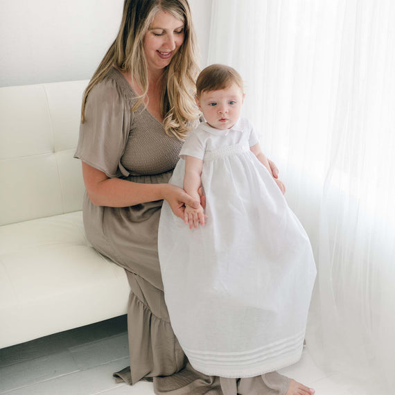 A woman with long hair in a brown dress sits on a white couch, holding a baby in an Oliver Convertible Gown | Romper & Skirt Set. They are both facing the camera near a bright window with white curtains.