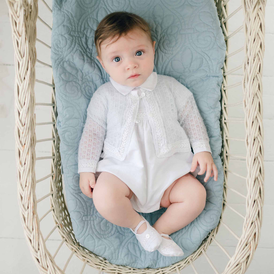 A baby with light brown hair wearing an Oliver Knit Sweater and light-colored shoes is lying in a wicker basket with a quilted blue cushion. The white outfit, adorned with delicate Venice lace trim, complements the baby's neutral expression as they gaze up.