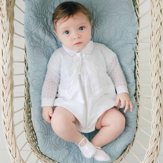 A baby with light brown hair wearing an Oliver Knit Sweater and light-colored shoes is lying in a wicker basket with a quilted blue cushion. The white outfit, adorned with delicate Venice lace trim, complements the baby's neutral expression as they gaze up.