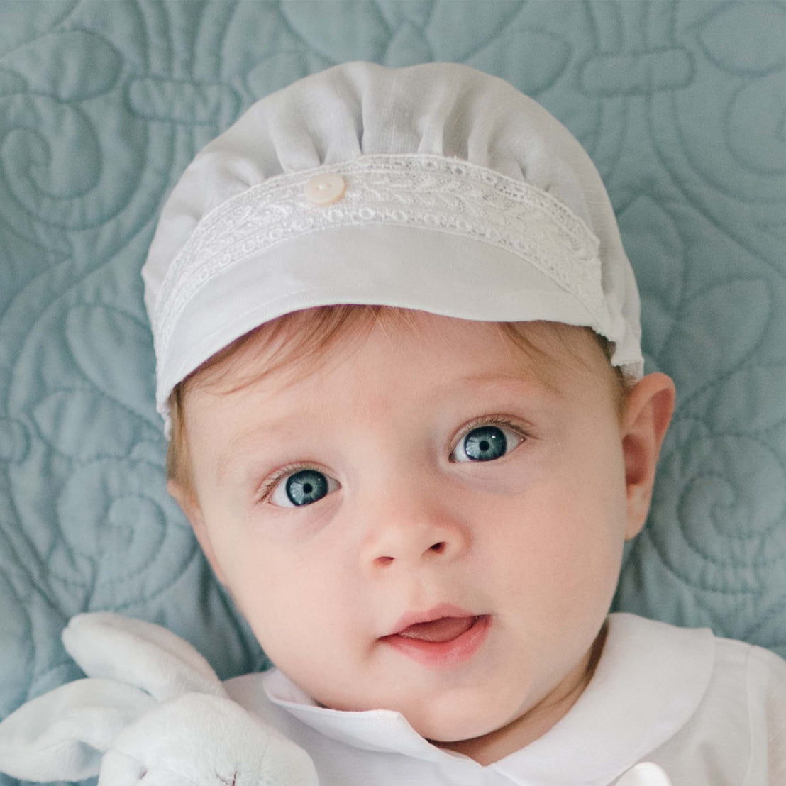 A baby with blue eyes wearing a white Oliver Linen Cap and white clothing is looking at the camera. The baby is lying on a light blue quilted surface. One of the baby's hands, partially visible, holds a small white plush toy adorned with delicate Venice lace.