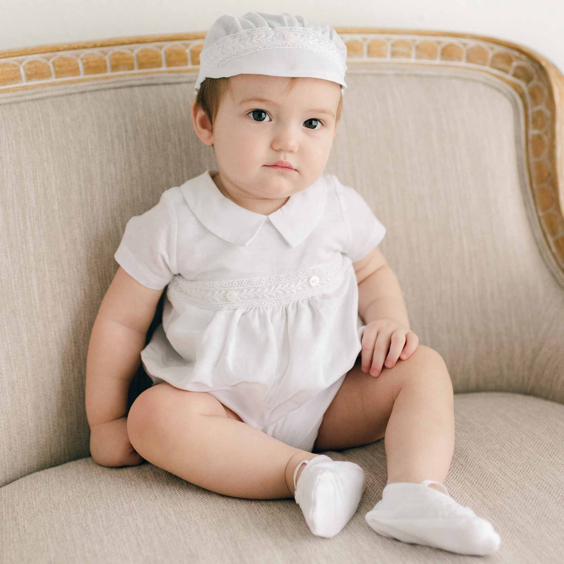 Smiling baby boy sitting on a chair and wearing the Oliver Bubble Romper (and matching Oliver Linen Cap).