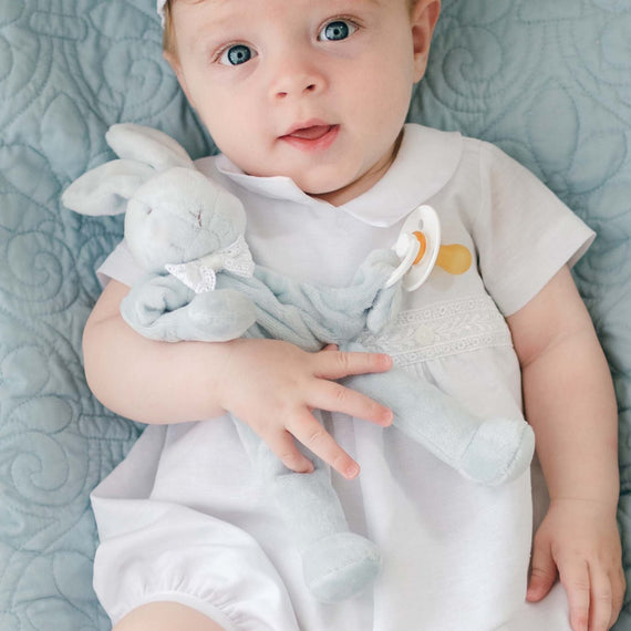 A baby with bright blue eyes lies on a soft, quilted blue blanket. The baby is wearing the Oliver Romper and matching linen cap in white, and is holding an Oliver Silly Bunny Buddy pacifier holder in light blue, made from soft velour. A baby-safe pacifier clip is attached to the bunny's hand. The baby looks up with a curious expression.