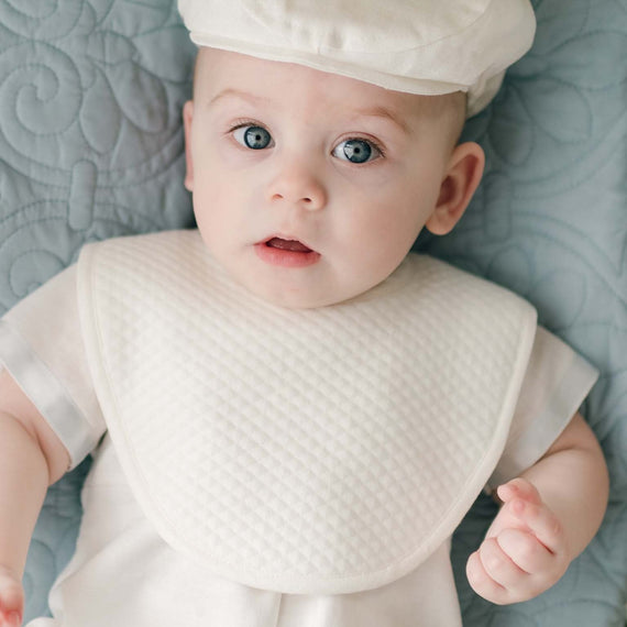 A baby wearing a white cap, white outfit with linen trim, and an Owen Christening Bib is lying on a light blue quilted blanket. The baby has wide blue eyes and a slight expression of surprise or curiosity. The setting appears to be indoors.