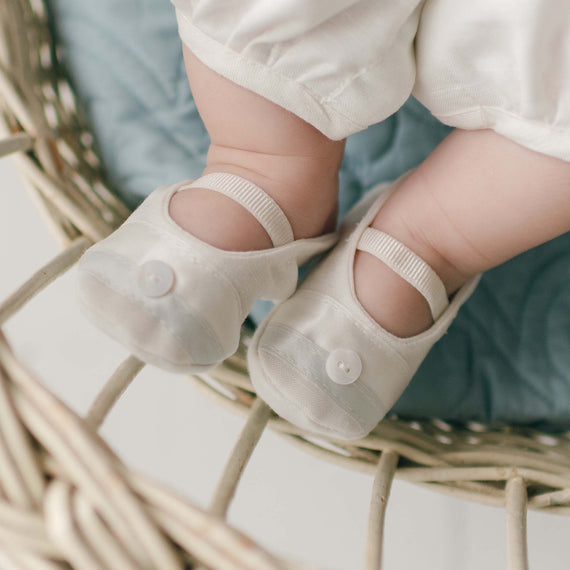 A baby's feet in Owen Linen Christening Booties with a button detail are resting in a woven wicker basket lined with a light blue quilted fabric. A delicate blue silk ribbon is tied around the basket's handle, adding a touch of charm. The baby is wearing white pants, and the image focuses on the lower legs and feet.
