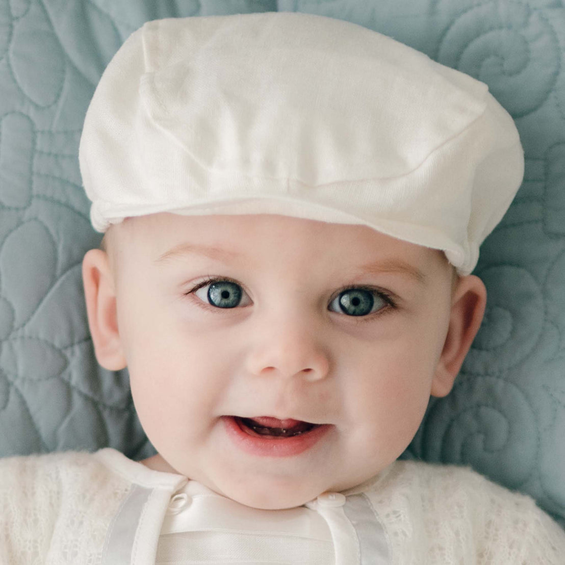 A baby with blue eyes is wearing a handmade white Oliver Linen Newsboy Cap and white clothing. The baby is looking directly at the camera with a slight smile, positioned against a quilted light blue background.
