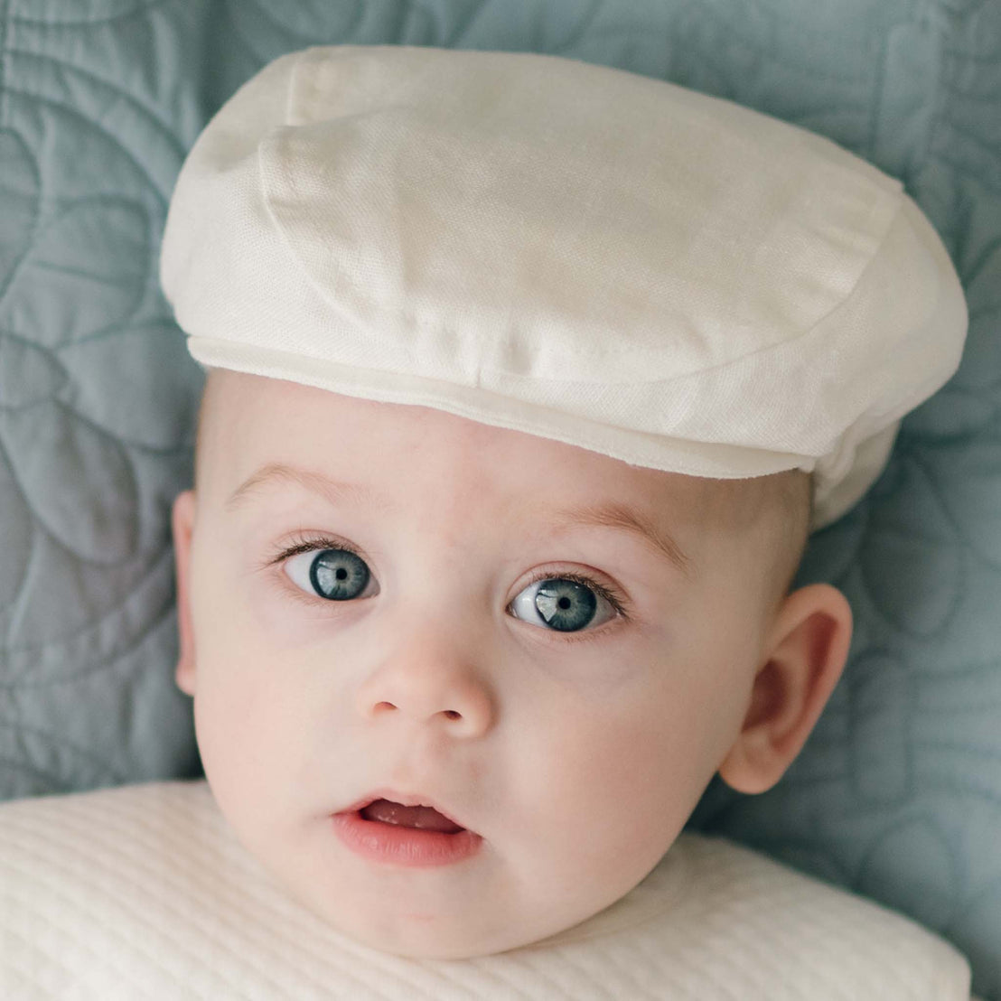 A baby with blue eyes is looking at the camera, wearing a handmade white christening outfit and an Oliver Linen Newsboy Cap. The background appears to be a textured fabric.