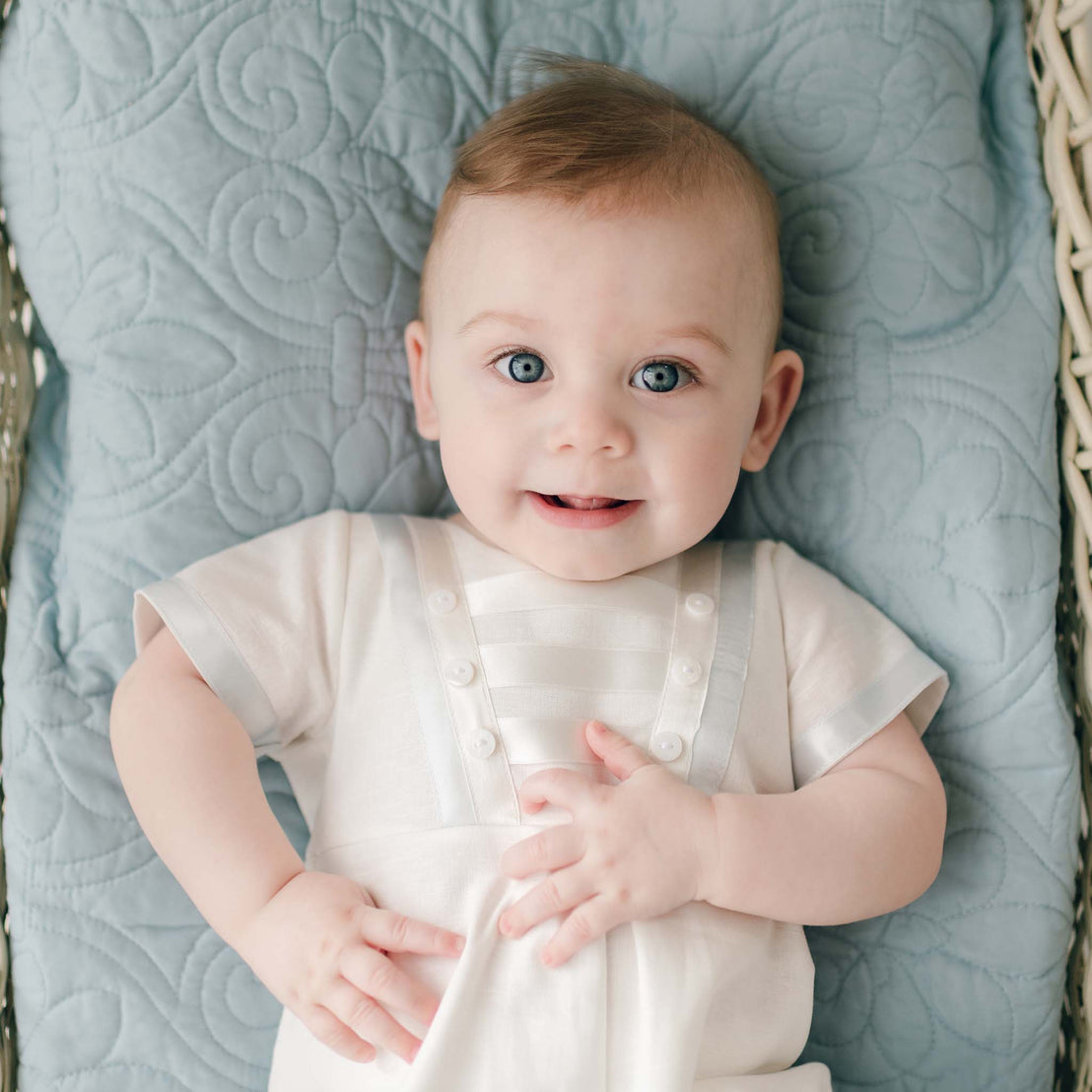 A baby with blue eyes and light brown hair is lying on a light blue quilted blanket. Clad in an Owen Linen Christening Romper, the baby looks up, smiling slightly, with one hand resting on the chest and the other on the blanket—perfect for his Christening day outfit.