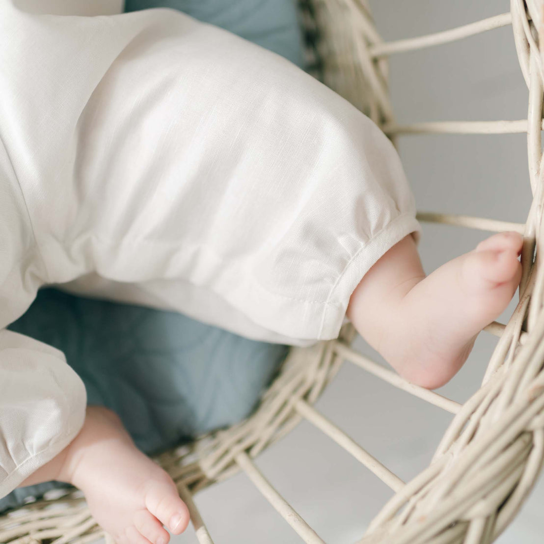 Close-up image of a baby’s legs and feet wearing an Owen Linen Christening Romper, sitting in a white wicker chair. The baby is resting on a blue cushion. Only the lower part of the baby's body is visible.