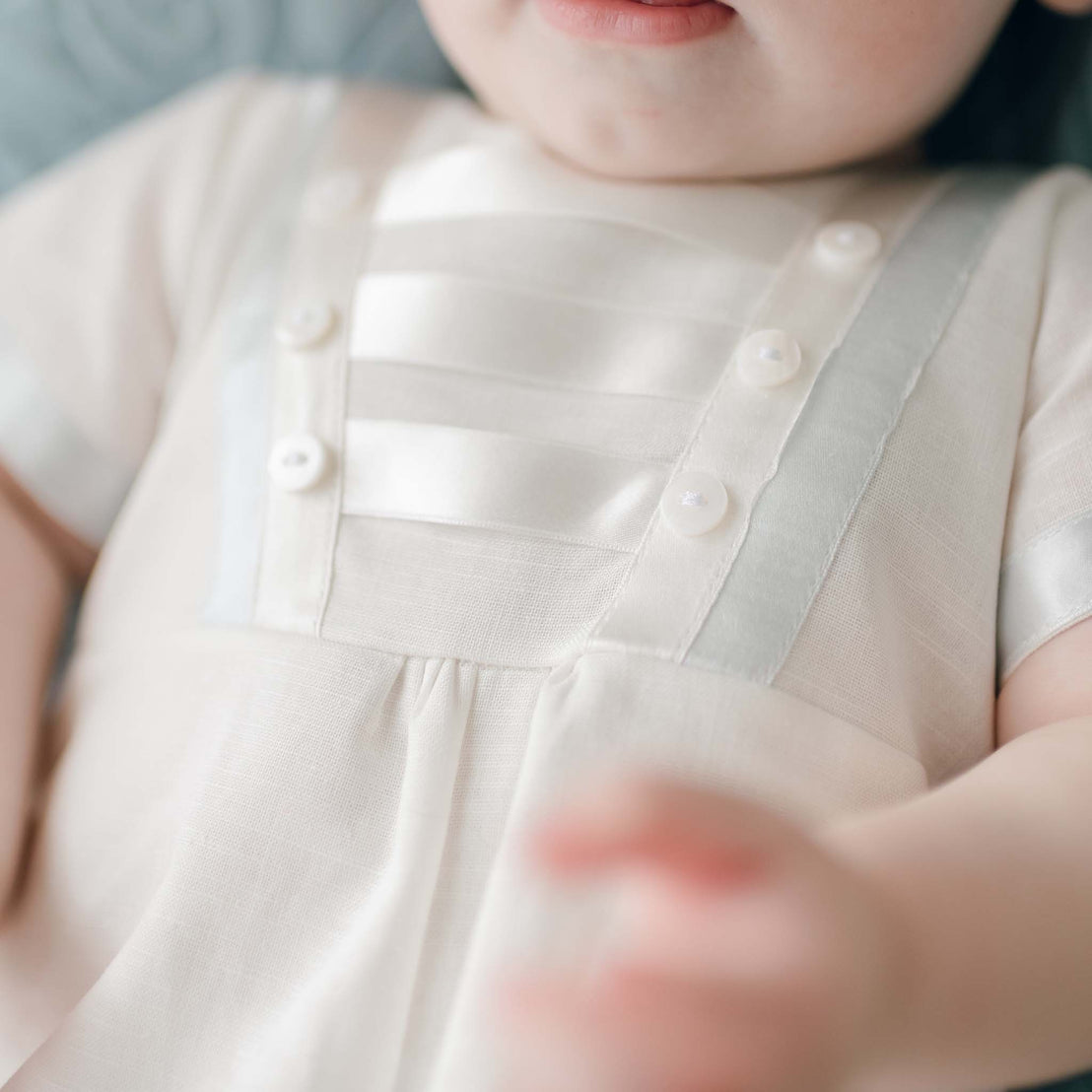 Close-up of a baby wearing an Owen Linen Christening Romper with satin stripes and small buttons. The focus is on the upper part of the outfit, akin to a Christening day outfit, and the baby's face is partially visible, showing the chin and lips. The baby's arm is out of focus in the foreground.