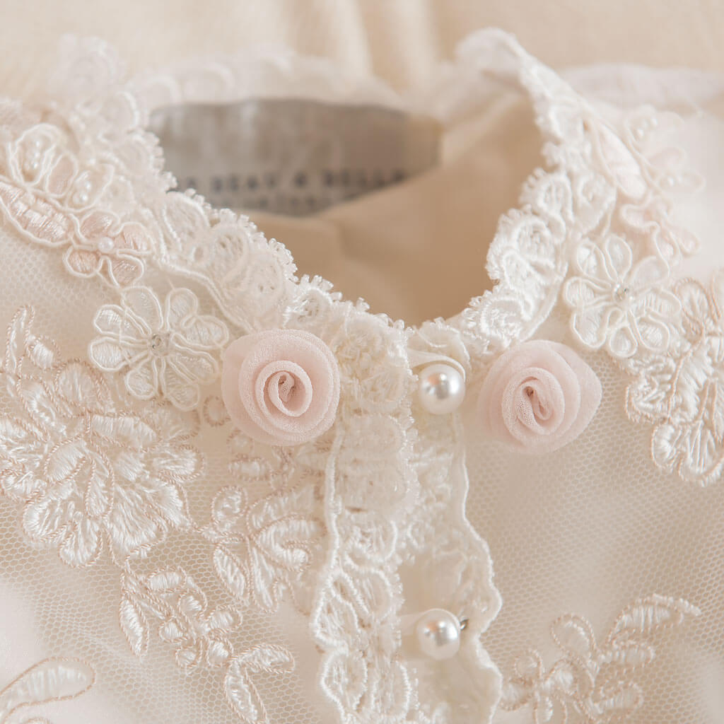 Close-up of a delicate lace garment with intricate floral embroidery. The fabric features two small pink rosettes and pearl-like buttons. The lace has a floral pattern and an elegant, vintage appearance.