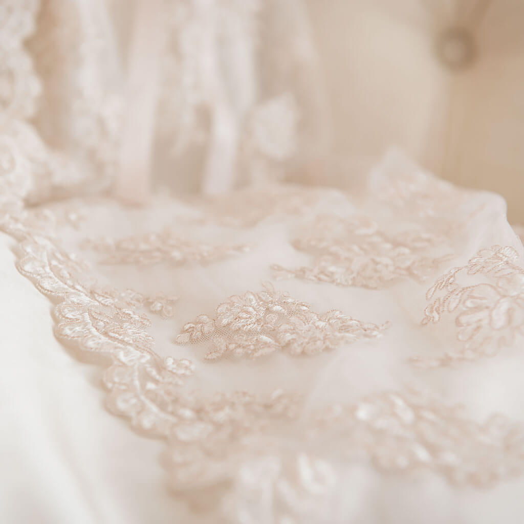 Close-up of delicate lace fabric in soft beige tones, adorned with intricate floral patterns, draped over a light, blurred background. The texture and detailing of the lace are in clear focus.
