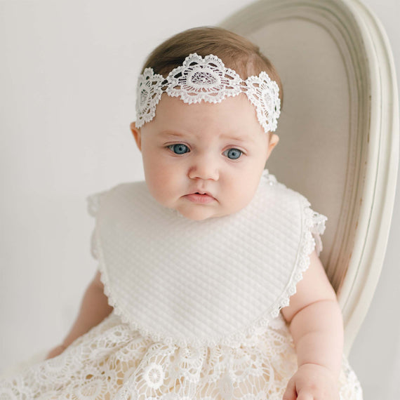 A baby wearing a white, floral lace headband and a Poppy Christening Bib with a lace dress sits on a beige cushioned chair. The baby has a neutral expression and blue eyes, looking slightly off to the side. The background is plain and light-colored.