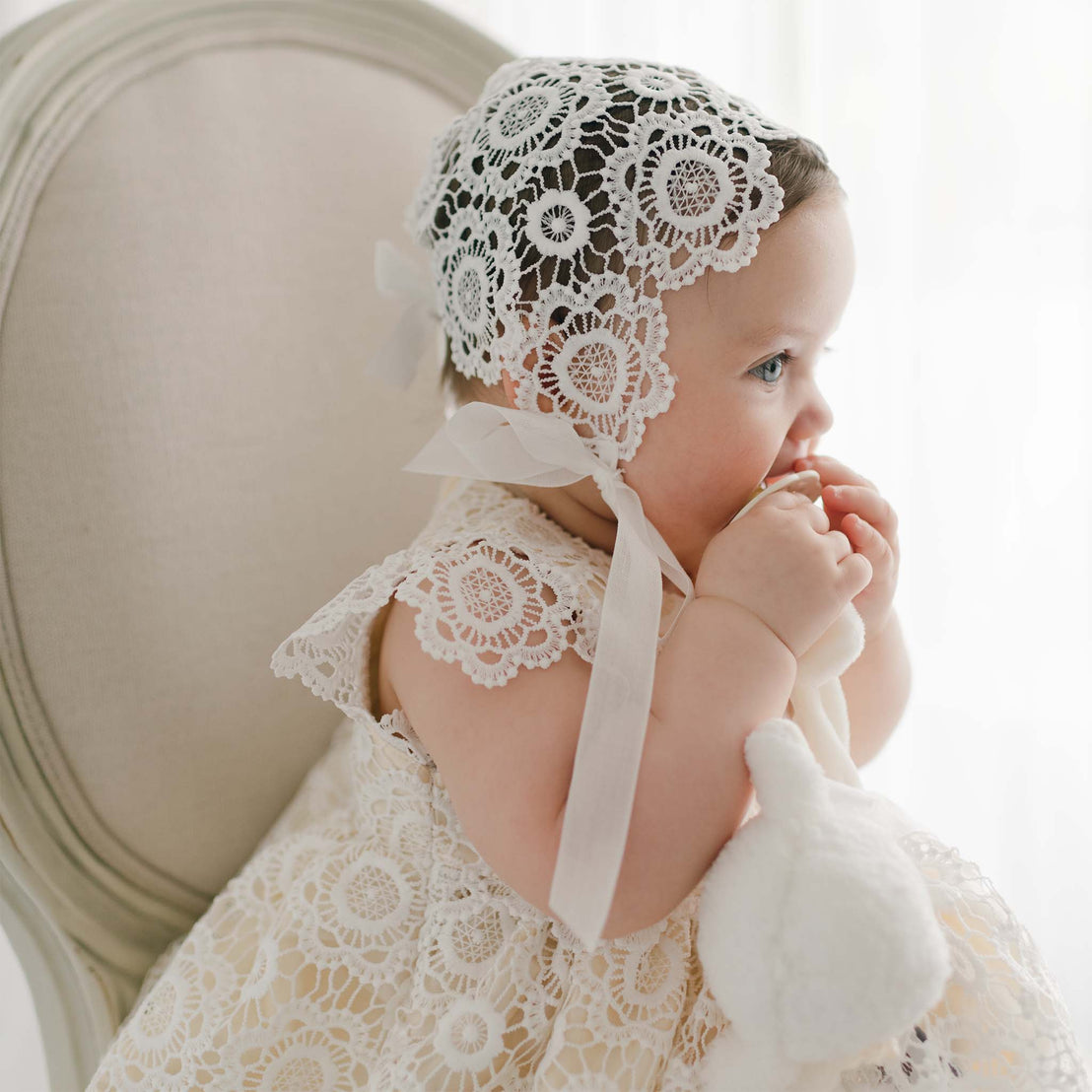 A baby sits on a chair wearing a white lace Poppy Blessing Dress & Bonnet. The baby is facing sideways, holding a white stuffed animal, and putting something in their mouth. The background is bright and softly lit.