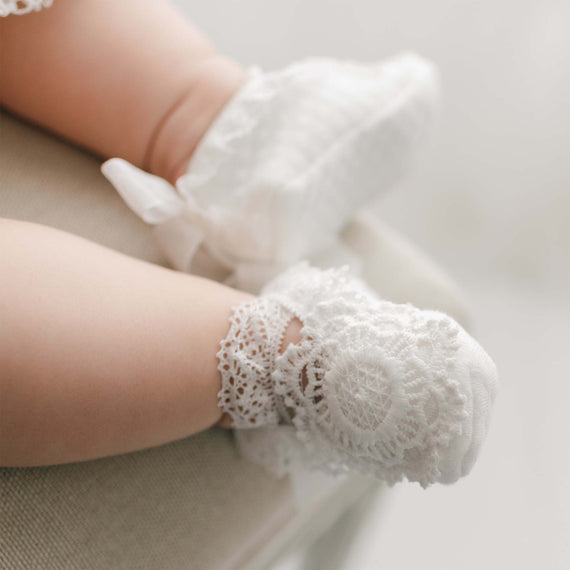 Close-up of a baby's legs and feet, wearing Poppy Christening Booties with intricate lace patterns. The baby is seated on a light-colored surface, with a soft, blurred background.