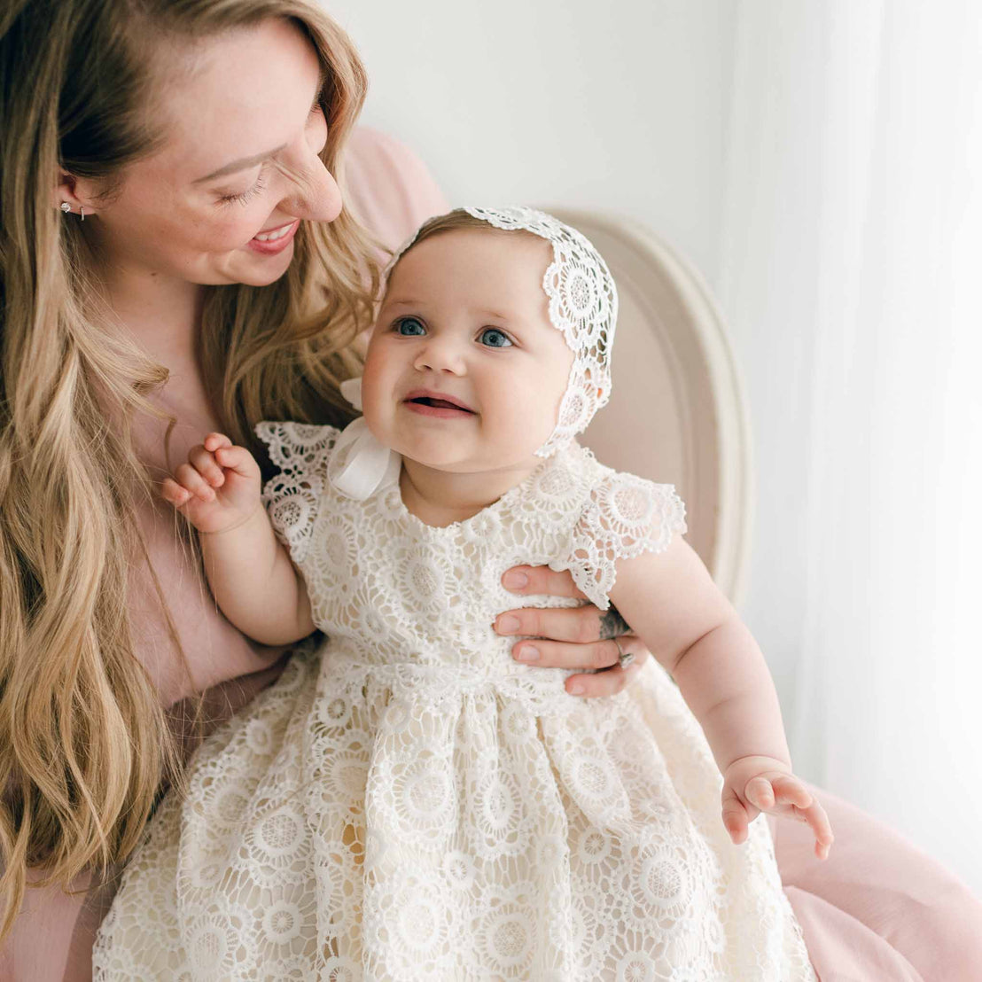 A woman with long blonde hair holds a baby dressed in a beautiful Poppy Christening Gown & Bonnet. Both are smiling warmly. They are indoors with a light background.