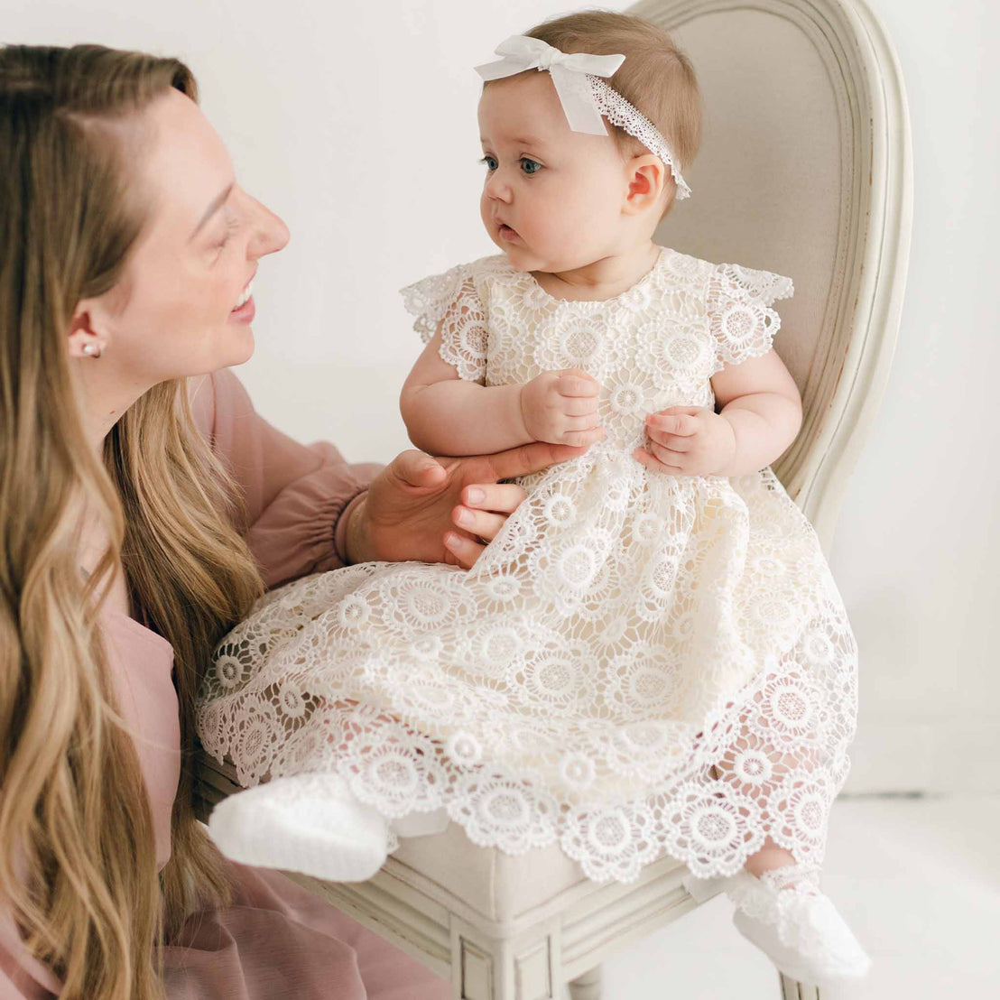 A woman with long, light brown hair is looking at a baby sitting on a white chair. The baby, dressed in a delicate Poppy Blessing Dress & Bonnet with intricate lace, headband, and white socks, is looking forward. The plain white backdrop enhances the soft and light ambiance of this special moment.