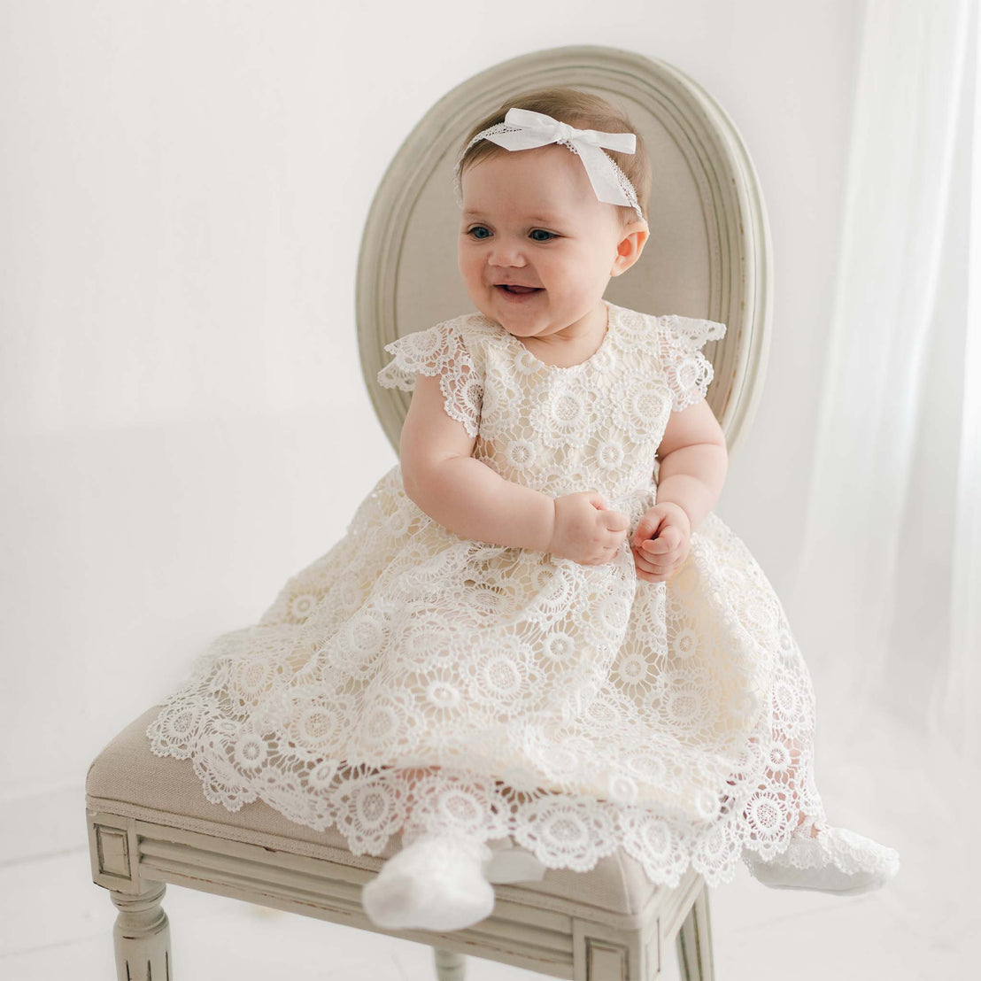 A baby sits on a cream-colored chair, wearing a beautiful Poppy Blessing Dress & Bonnet with lace sleeves and a white headband. The baby smiles and clasps hands together, with white socks on their feet. The background is a softly lit, plain white room.
