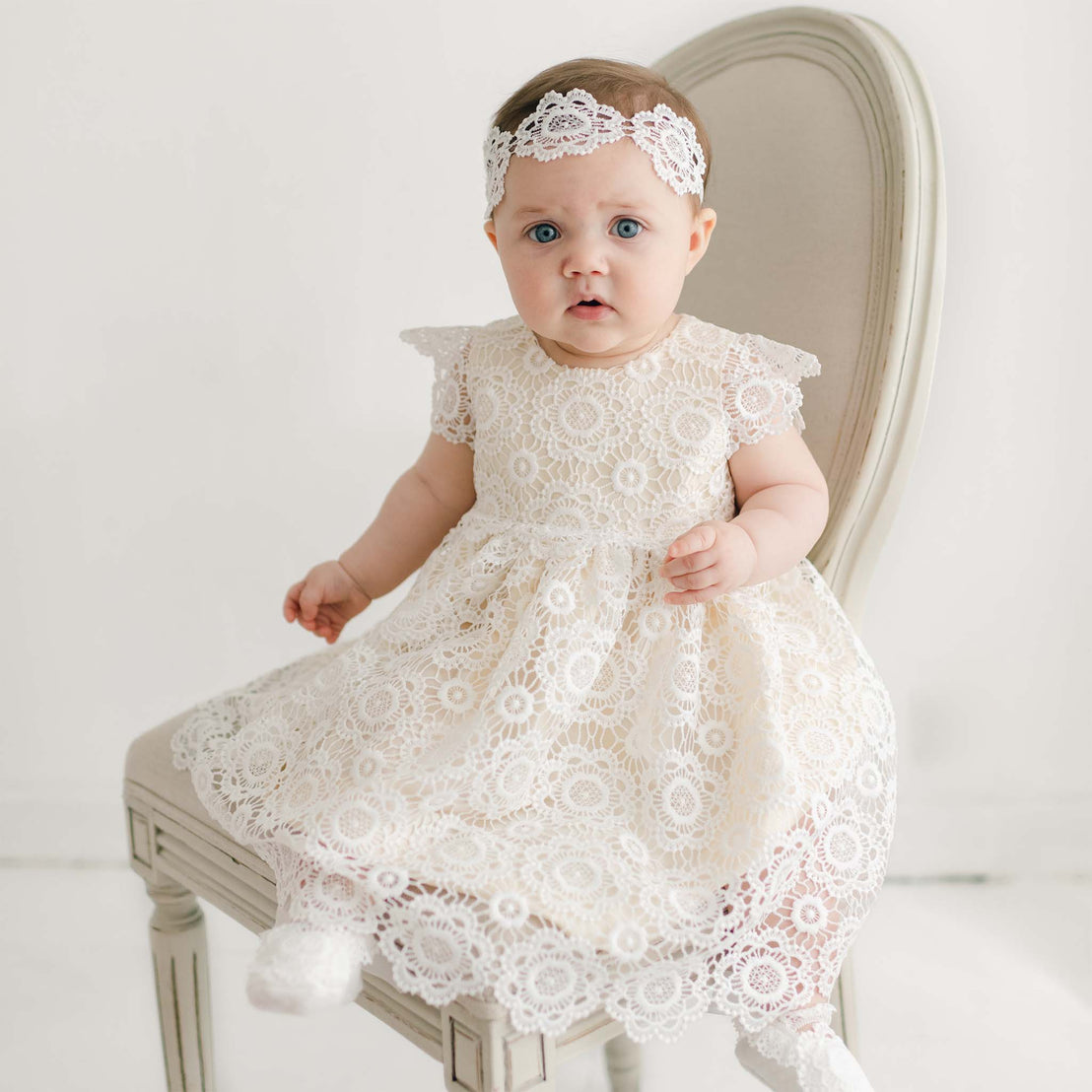 A baby sits on a beige chair, wearing a Poppy Blessing Dress & Bonnet with a silk dupioni lining. The baby has blue eyes and dark hair, looking directly at the camera with a neutral expression. The background is plain and white.