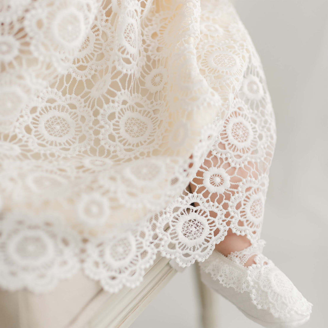 A close-up of a delicate Poppy Blessing Dress & Bonnet and matching shoes worn by an infant. The lace features intricate circular patterns with floral motifs, and the baby is seated on a white surface. The soft lighting highlights the texture and detail of the lace fabric, complemented by a silk dupioni lining.