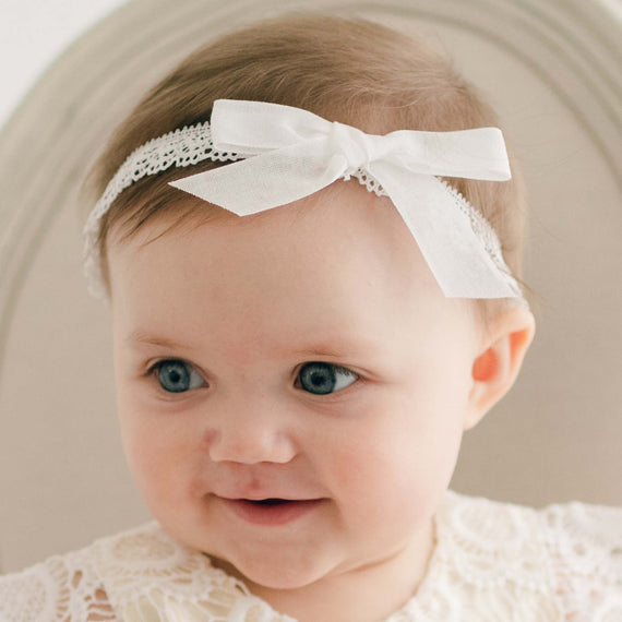 A baby with short brown hair wears a Poppy Christening Headband with a bow. The baby is gazing slightly to the side with a slight smile and is dressed in a white lace outfit. The background features a neutral-colored chair.