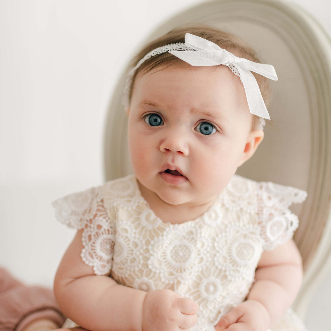 A baby with blue eyes and light brown hair sits on a chair. The baby is wearing a Poppy Blessing Dress & Bonnet adorned with a silk dupioni lining and has a white bow headband. The baby is looking slightly to the side with a neutral expression.
