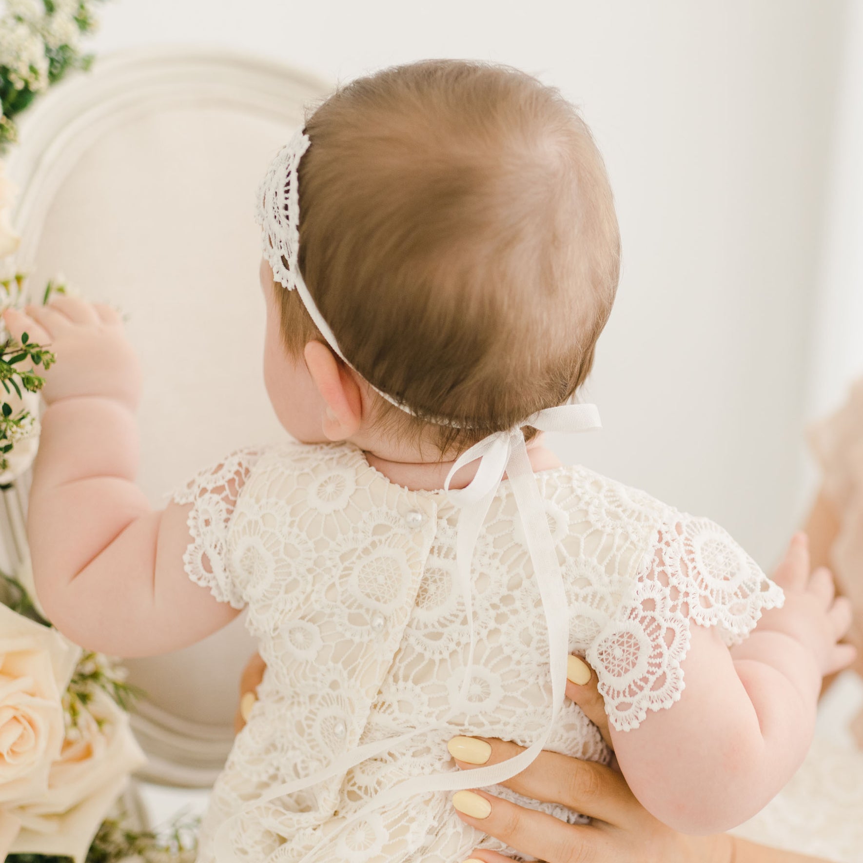 Back view of a baby wearing the Poppy Lace Headband, tied with a delicate white ribbon, and the coordinating lace gown with intricate floral patterns and pearl buttons.