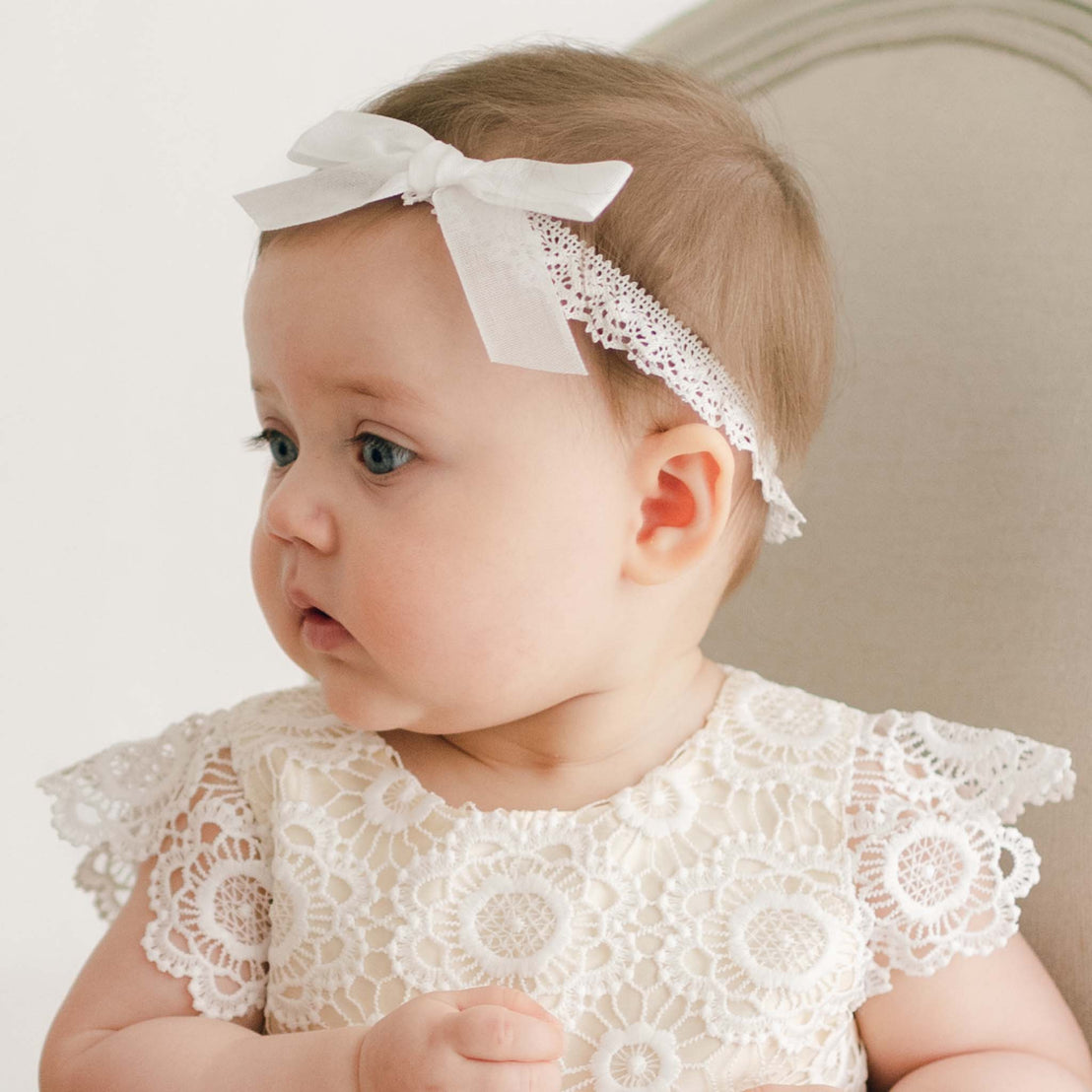 A baby seated on an off-white chair is wearing a white lace dress with floral patterns and a Poppy Christening Headband adorned with a bow. The baby is looking to the side with a neutral expression.