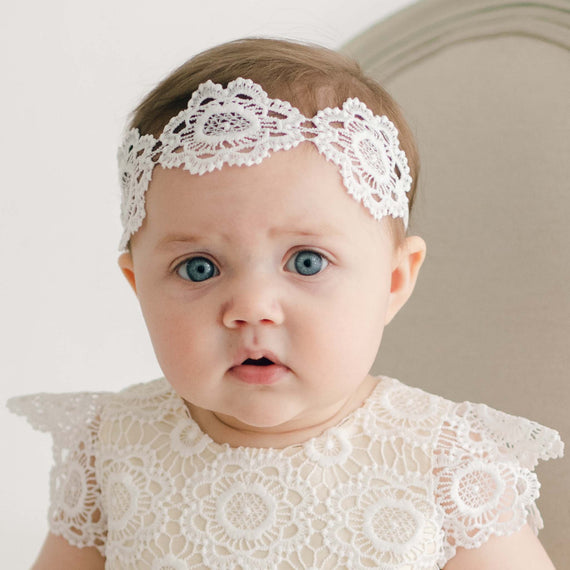 A baby with blue eyes wears a light ivory Poppy Lace Headband and a matching lace outfit. The baby is seated in front of a beige chair and looks towards the camera with a neutral expression. The background is plain and simple.