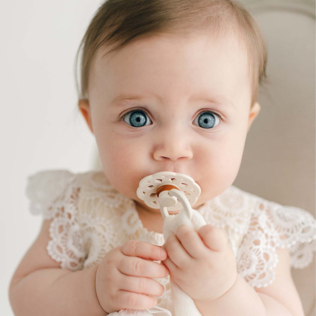 A baby with blue eyes is wearing a white lace outfit and holding a pacifier in their mouth with both hands, secured by a sweet Poppy Silly Lamb Buddy | Pacifier Holder. The background is plain and light-colored.