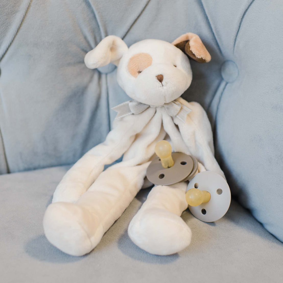 The Ezra Tan Silly Puppy Buddy stuffed animal sitting on a couch. This stuffed animal serves as a pacifier holder