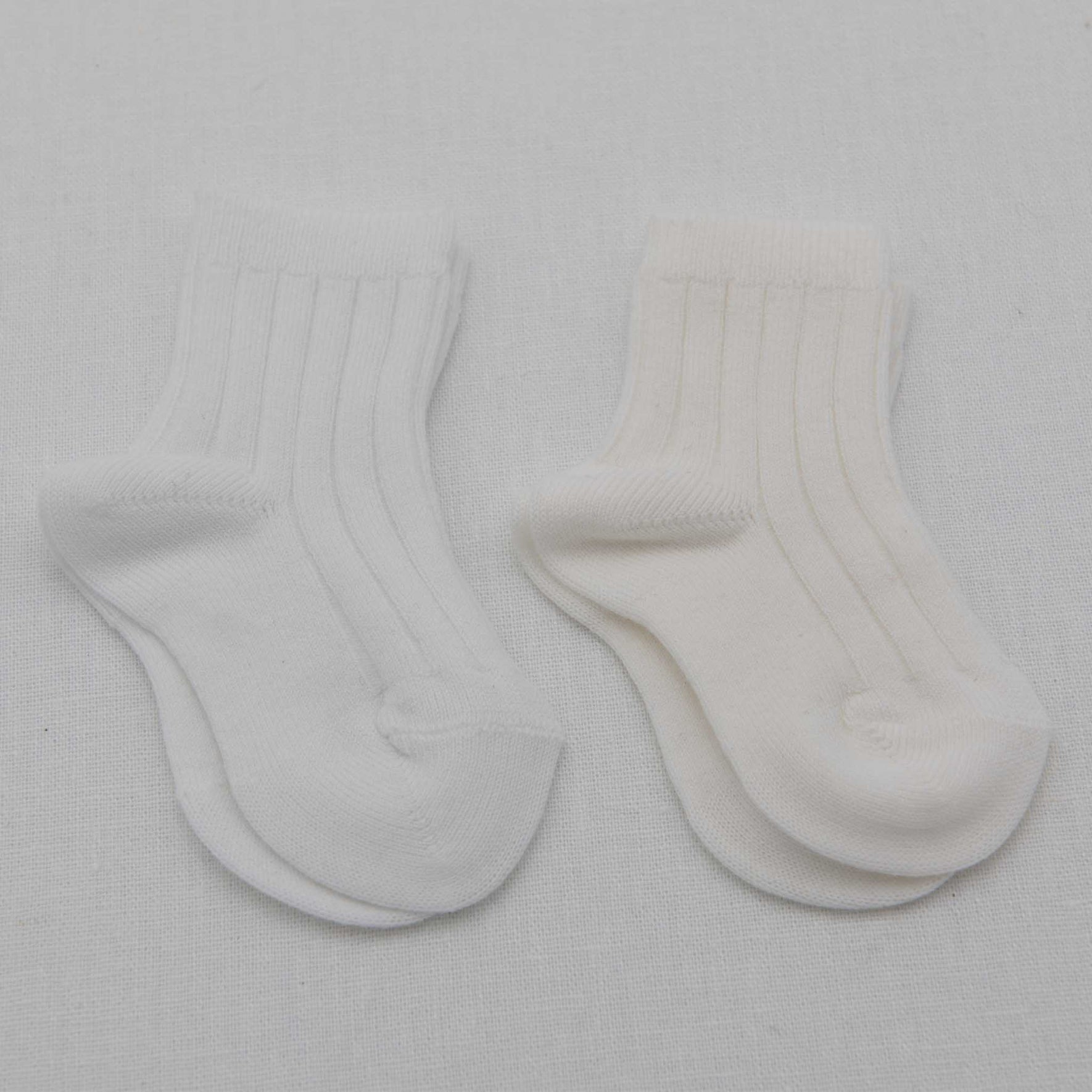Two pairs of Ribbed Socks are displayed on a white surface. Made in Spain, one pair features a light grey cotton blend and the other is white. Both pairs exhibit a simple, classic design with an enduring ribbed pattern.