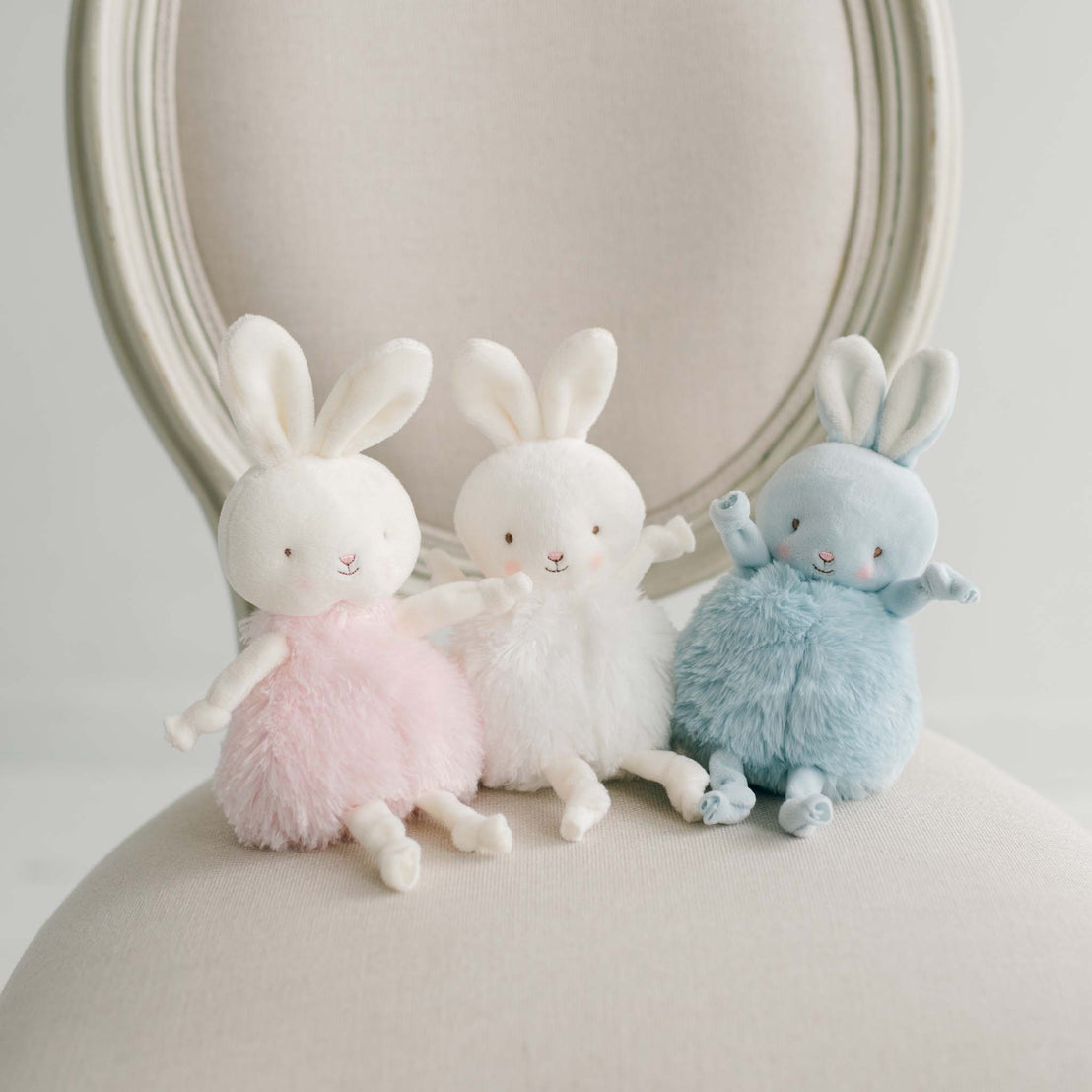 Three plush bunny toys in pink, white, and blue on a beige chair with a neutral background