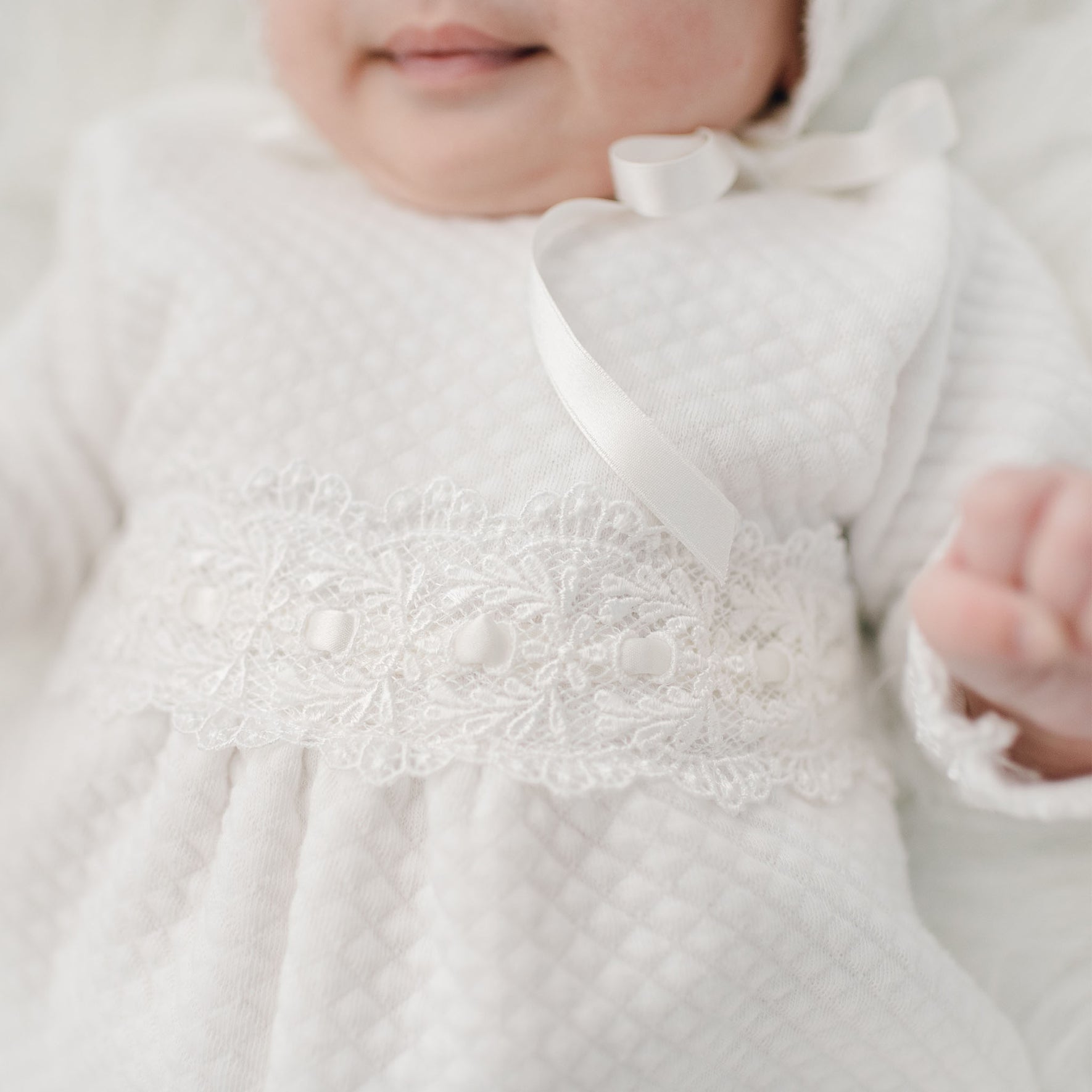 A close-up image of an infant wearing a white, quilted cotton outfit adorned with lace and ribbon details. The baby’s face is partially visible, and one hand is raised and slightly blurred in the foreground. The focus is on the intricate lace and ribbon on the Madeline Quilted Newborn Romper.