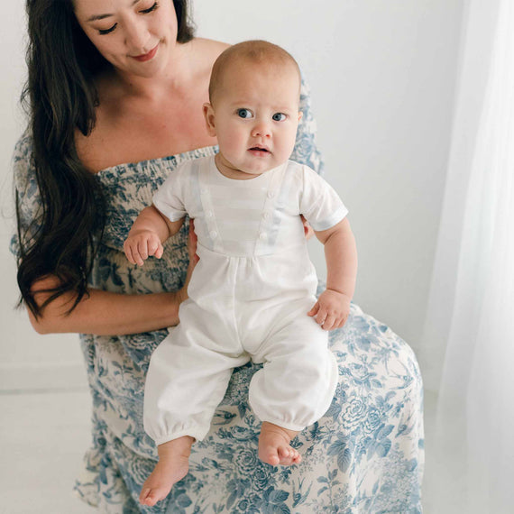 Baby boy sitting on his mother's lap and wearing the Owen Linen Romper featuring silk ribbon in ivory and light blue across the front bodice and sleeves