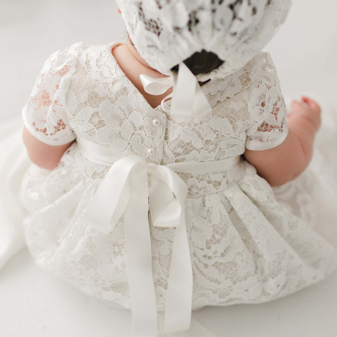 A baby dressed in a Rose Christening Gown & Bonnet, seen from behind. The dress is detailed with a silk ribbon at the back. The setting is a bright room with a soft ambiance.