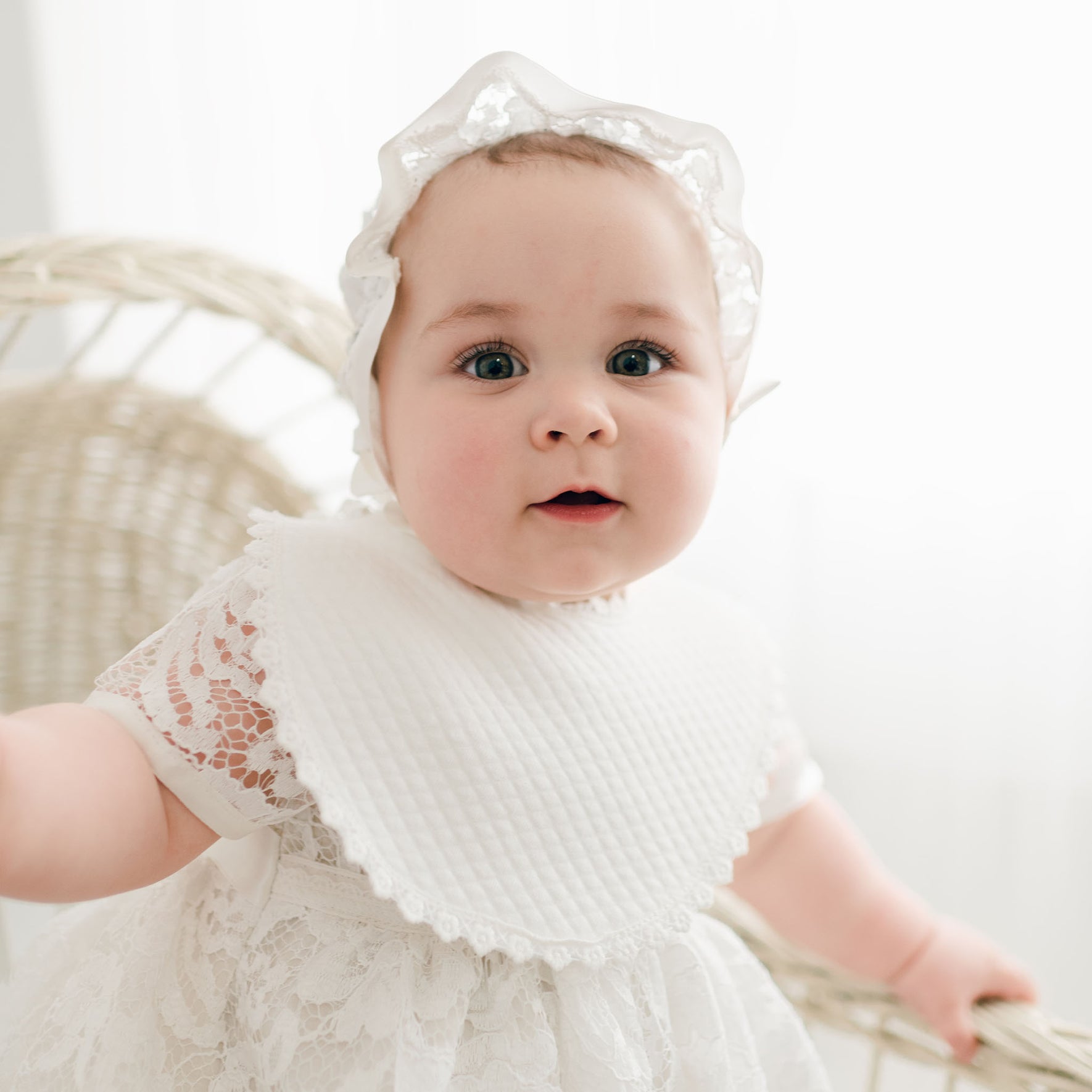 A baby with wide blue eyes and a white headband sits in a rattan chair, wearing an ivory quilted cotton dress and a Rose Christening Bib, looking directly at the camera with a curious expression.