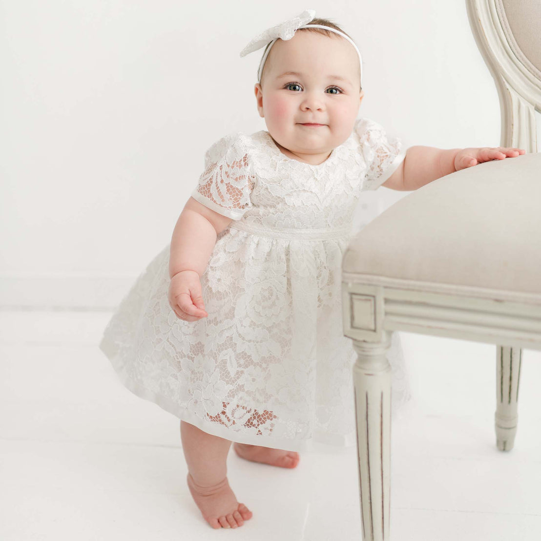 A baby girl in the Rose Romper Dress and matching Bow Headband stands barefoot, leaning on a vintage chair in a bright, white studio setting.