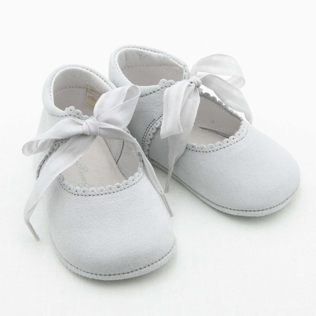 A pair of Emily Suede Tie Mary Janes, featuring a light gray color, scalloped edges, and white ribbon tie closures are showcased against a white background. These handmade shoes appear soft and delicate, making them perfect for an infant.