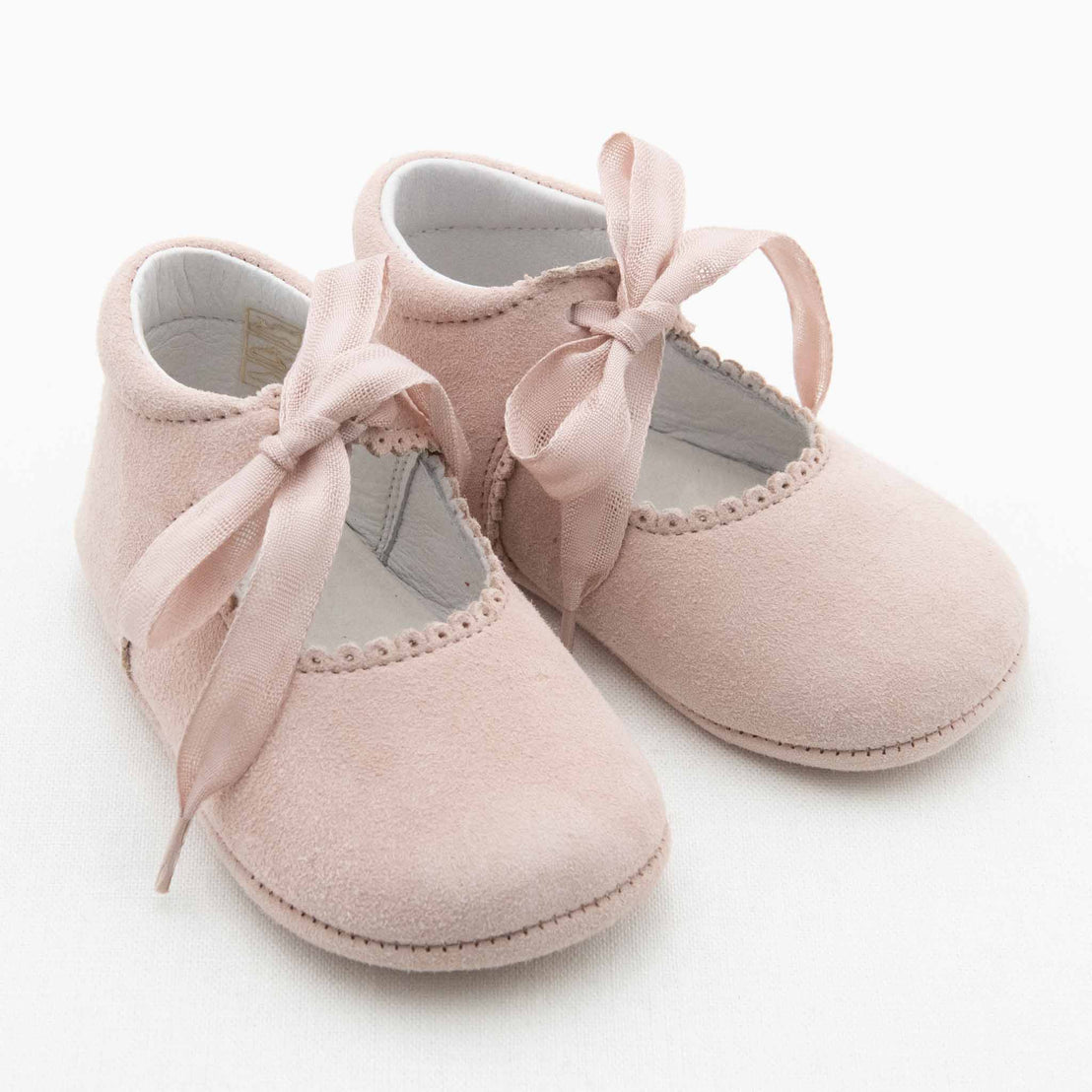 A pair of soft pink June Suede Tie Mary Janes with decorative stitching around the opening and ribbon laces tied in bows. This handmade baby footwear has a suede-like texture and is designed for infants. They are placed on a plain white background.