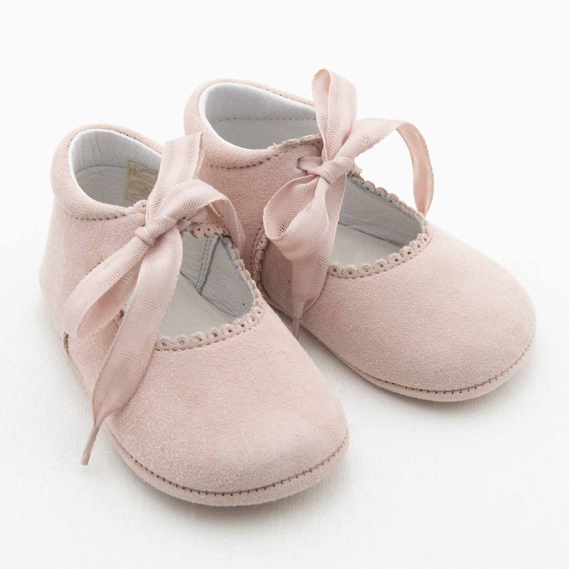 The Thea Suede Tie Mary Janes by Baby Beau & Belle are a pair of light pink handmade baby shoes featuring a velvet texture and satin ribbons. These shoes have scalloped edges, a soft sole, and are designed for infants. They are styled as Mary Janes and photographed against a white background.