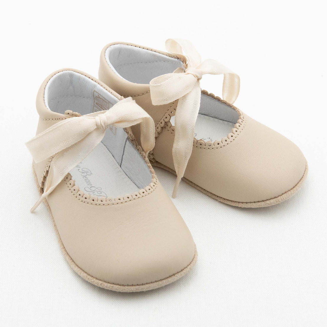 A pair of Tan Tie Mary Janes with satin ribbon ties and scalloped trim rests on a white surface. These handmade beige baby shoes feature a soft, rounded toe and a simple, elegant design. The interior lining is crisp white, showcasing the quality typical of Baby Beau & Belle.