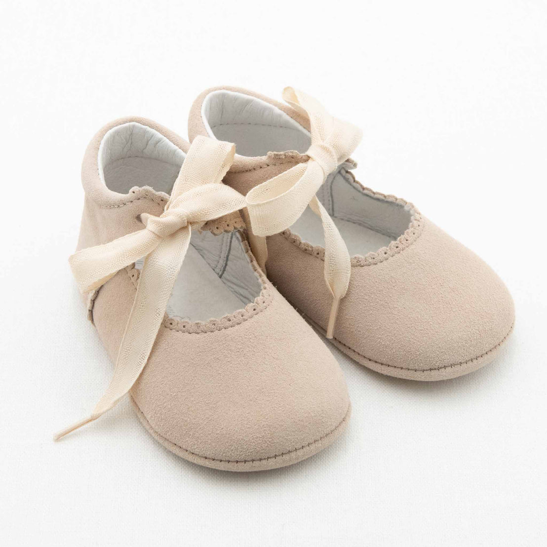 A pair of light beige Emily Suede Tie Mary Janes, handmade by Baby Beau & Belle, featuring soft fabric with ribbon ties and scalloped edges. Placed on a white background, these adorable baby shoes have cushioned interiors and flat soles for ultimate comfort.